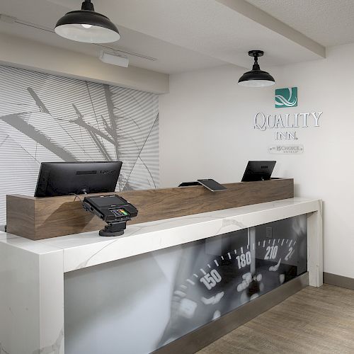 The image shows a modern, minimalist hotel reception desk with two computers, card readers, and a decorative wall with a logo and large graphic.