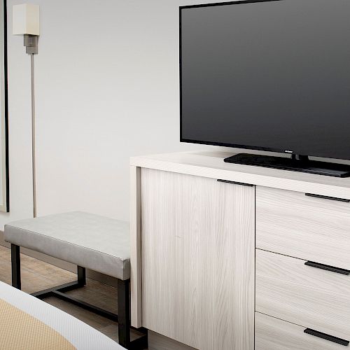 A modern hotel room features a flat-screen TV on a light wood dresser, a bench, a mirror, and a portion of a bed with a striped cover visible.