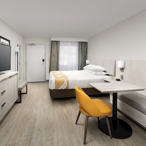 A modern hotel room with a bed, flat-screen TV, mini-fridge, small table and chairs, and a window with curtains.