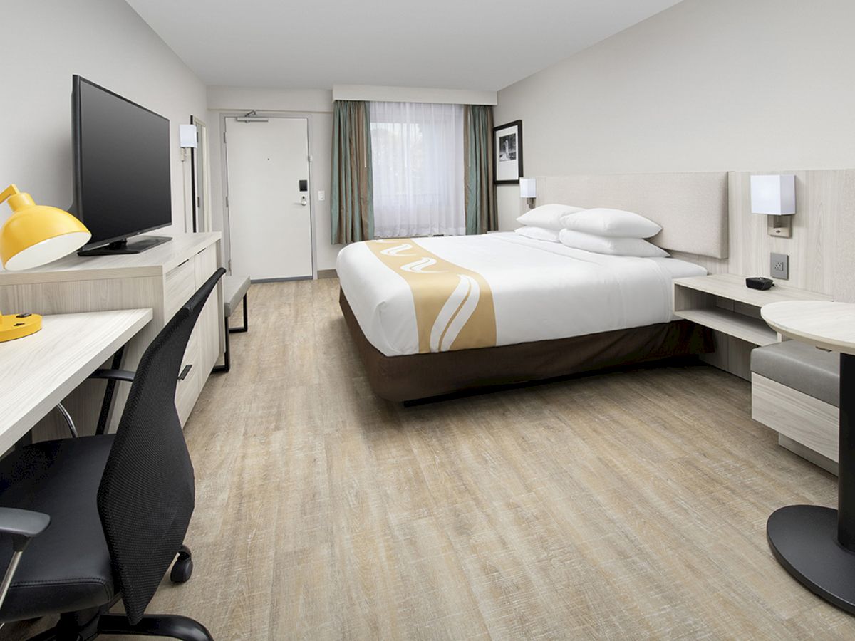 A modern hotel room with a double bed, work desk, yellow lamp, flat-screen TV, chair, small round table, and window with curtains at the far end.