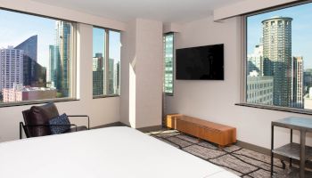 A hotel room with large windows offering a city view, a flat-screen TV, a white bed, a chair, and a wooden cabinet.