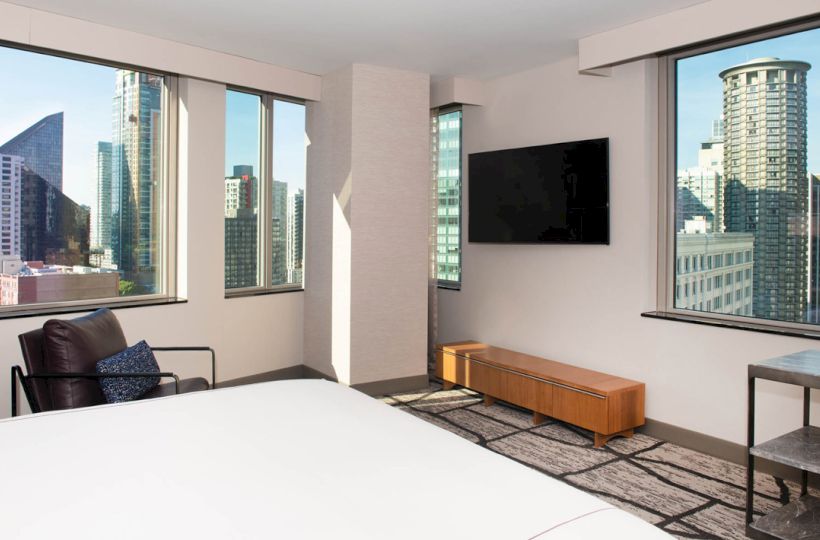 A hotel room with large windows offering a city view, a flat-screen TV, a white bed, a chair, and a wooden cabinet.