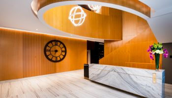 A modern lobby with a marble reception desk, wooden walls, a large wall clock, unique ceiling design, and flowers on the counter ends the sentence.