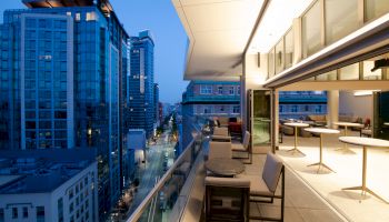 A modern city scene with tall buildings during dusk, and a spacious, furnished balcony of a high-rise apartment or office overlooking a lit street.