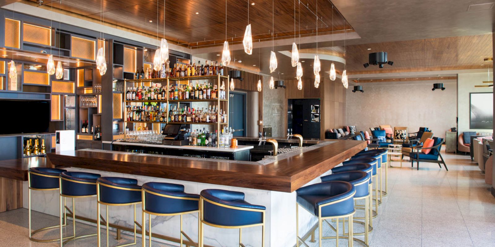 A modern bar with a wooden counter, stylish chairs, various liquors on display, pendant lights, and a lounge area with chairs and sofas in the background.
