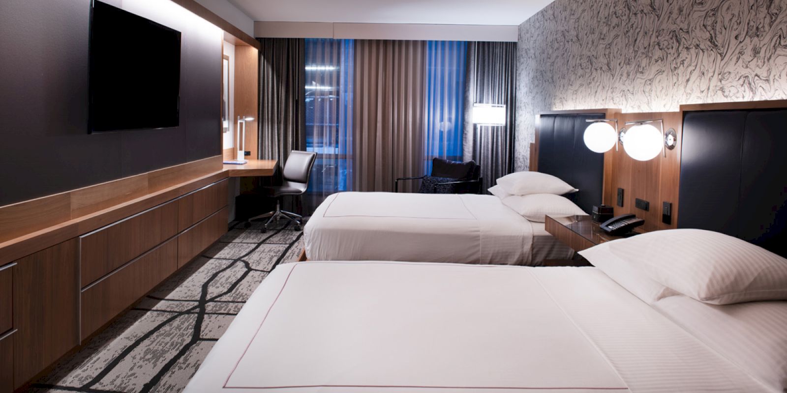 A modern hotel room features two neatly made beds, a wall-mounted TV, desk, chair, and lamps, with curtains covering large windows.
