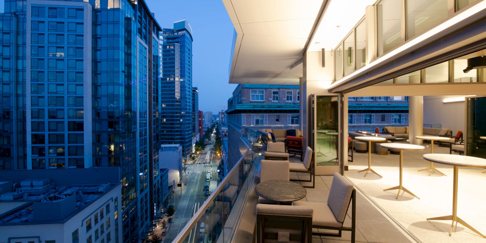 A modern balcony with tables and chairs overlooks a city street lined with high-rise buildings during twilight.