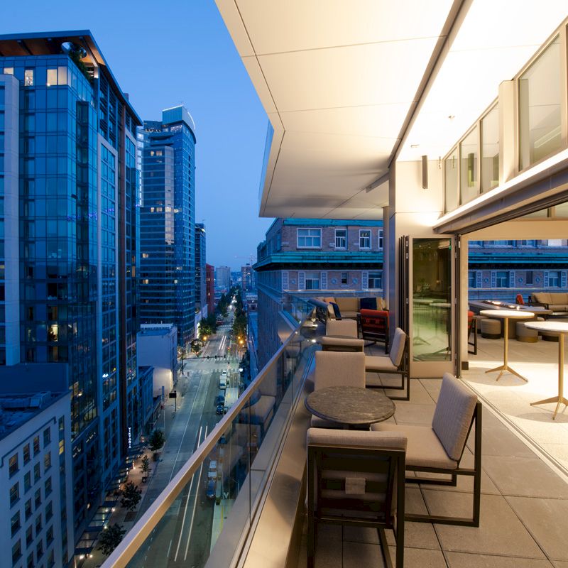 A modern balcony overlooks an urban skyline at dusk, with lounge chairs and tables near large windows of a building.