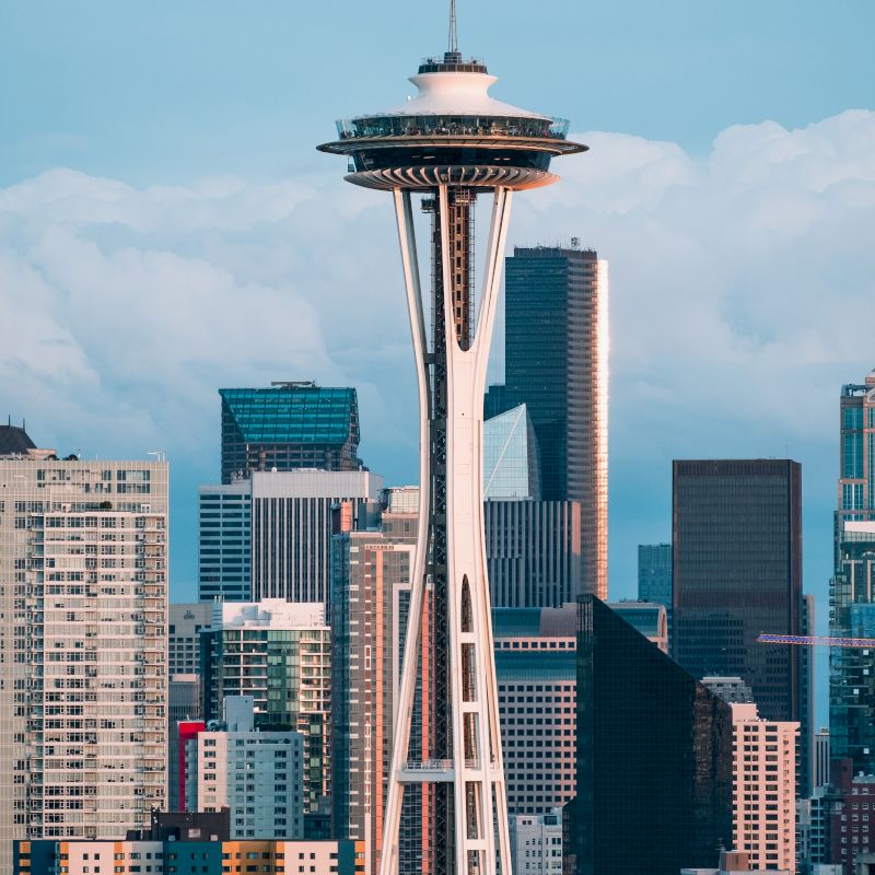 The image shows the Space Needle, a famous landmark, with the skyline of downtown Seattle in the background under a partly cloudy sky.