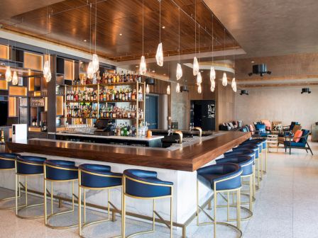 A modern bar with a variety of liquor bottles, stylish bar stools, hanging lights, and a seating area in the background.