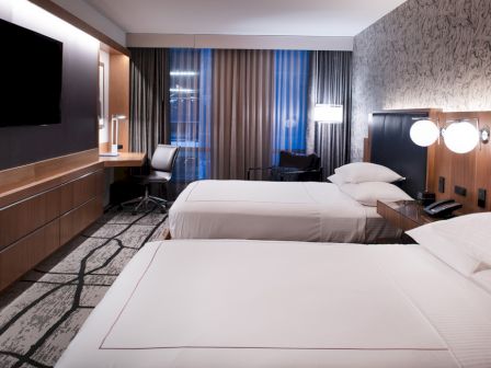 A modern hotel room with two double beds, a wall-mounted TV, a desk with a chair, and contemporary decor featuring patterned wallpaper.