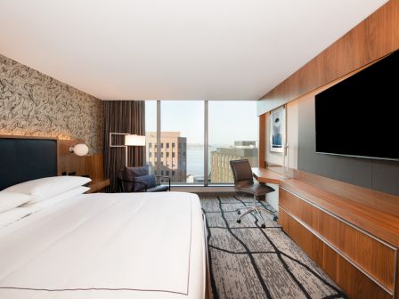 The image shows a modern hotel room with a large bed, a desk, a chair, a flat-screen TV, and a large window with a city view.