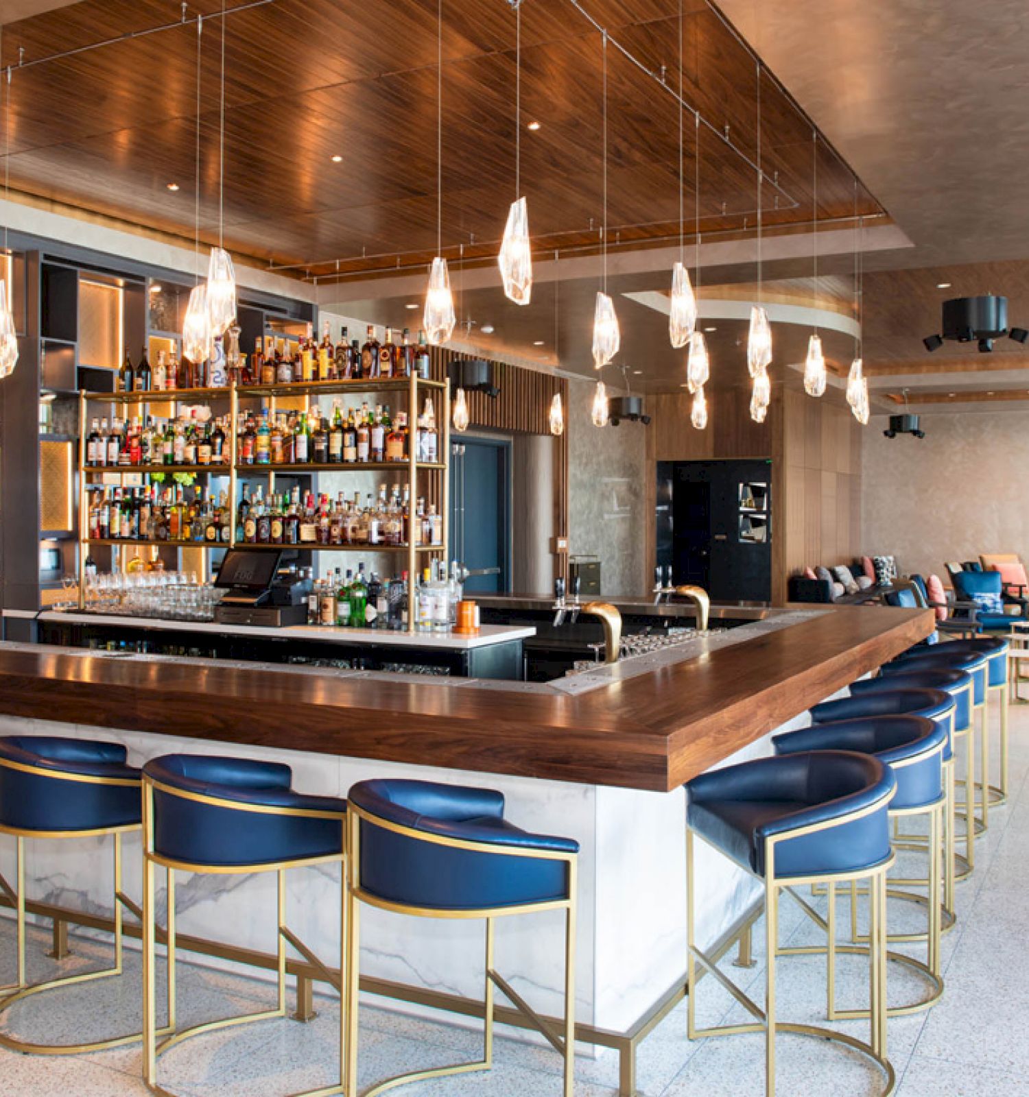This image shows a modern bar with blue seats, a wooden countertop, and various bottles displayed on shelves. Pendant lights hang from the ceiling.