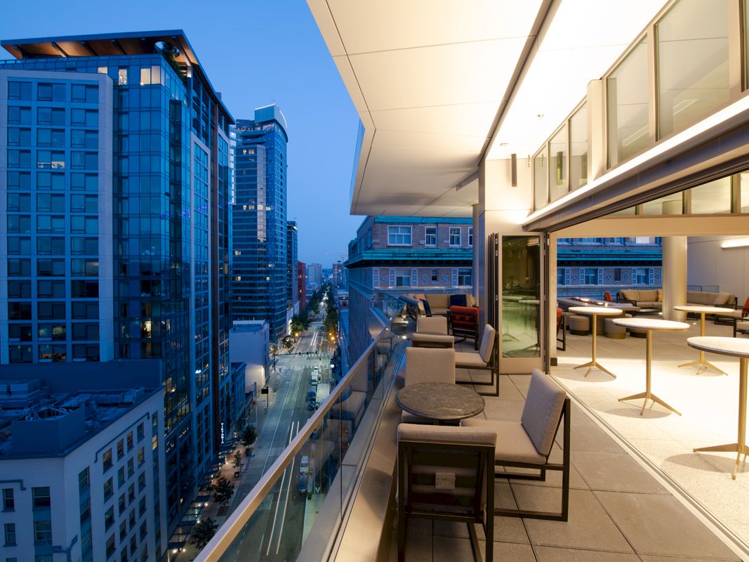 This image shows a modern cityscape with tall buildings and a balcony with seating and tables overlooking the street below.