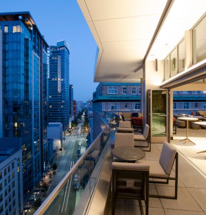 A modern cityscape at dusk with high-rise buildings and a stylish balcony featuring seating areas and tables overlooking the street below.