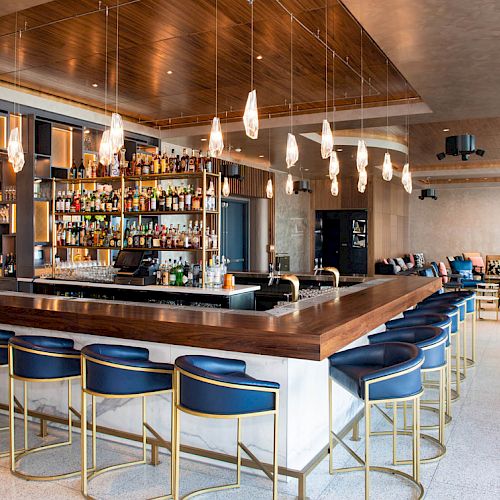 A modern bar with a wooden counter, upholstered stools, various liquor bottles on shelves, hanging lights, and a cozy seating area in the back.