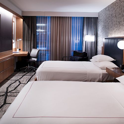 The image shows a modern hotel room with two double beds, a wall-mounted TV, a desk with a chair, and a large window with curtains.