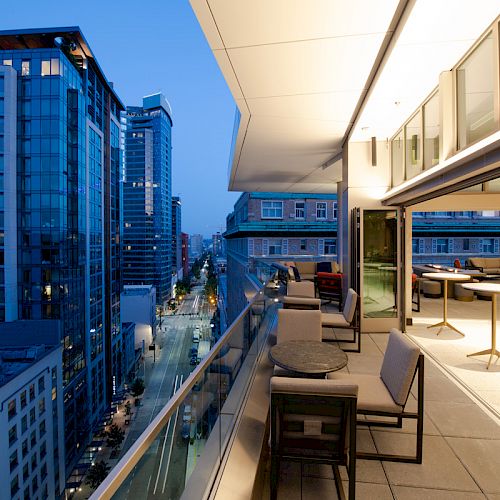 A modern balcony overlooking a cityscape at dusk, with seating and tables, adjacent to a well-lit indoor space with more tables and chairs.