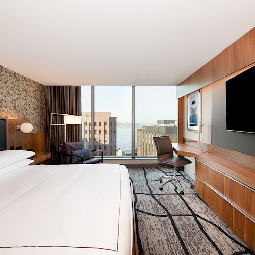 This image shows a modern hotel room with a large bed, a wall-mounted TV, a desk, and a window offering a cityscape view.