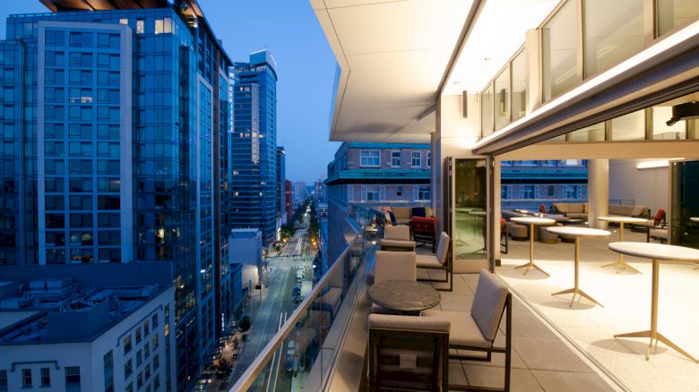 The image shows a cityscape with high-rise buildings and an outdoor balcony area furnished with tables and chairs in the evening.