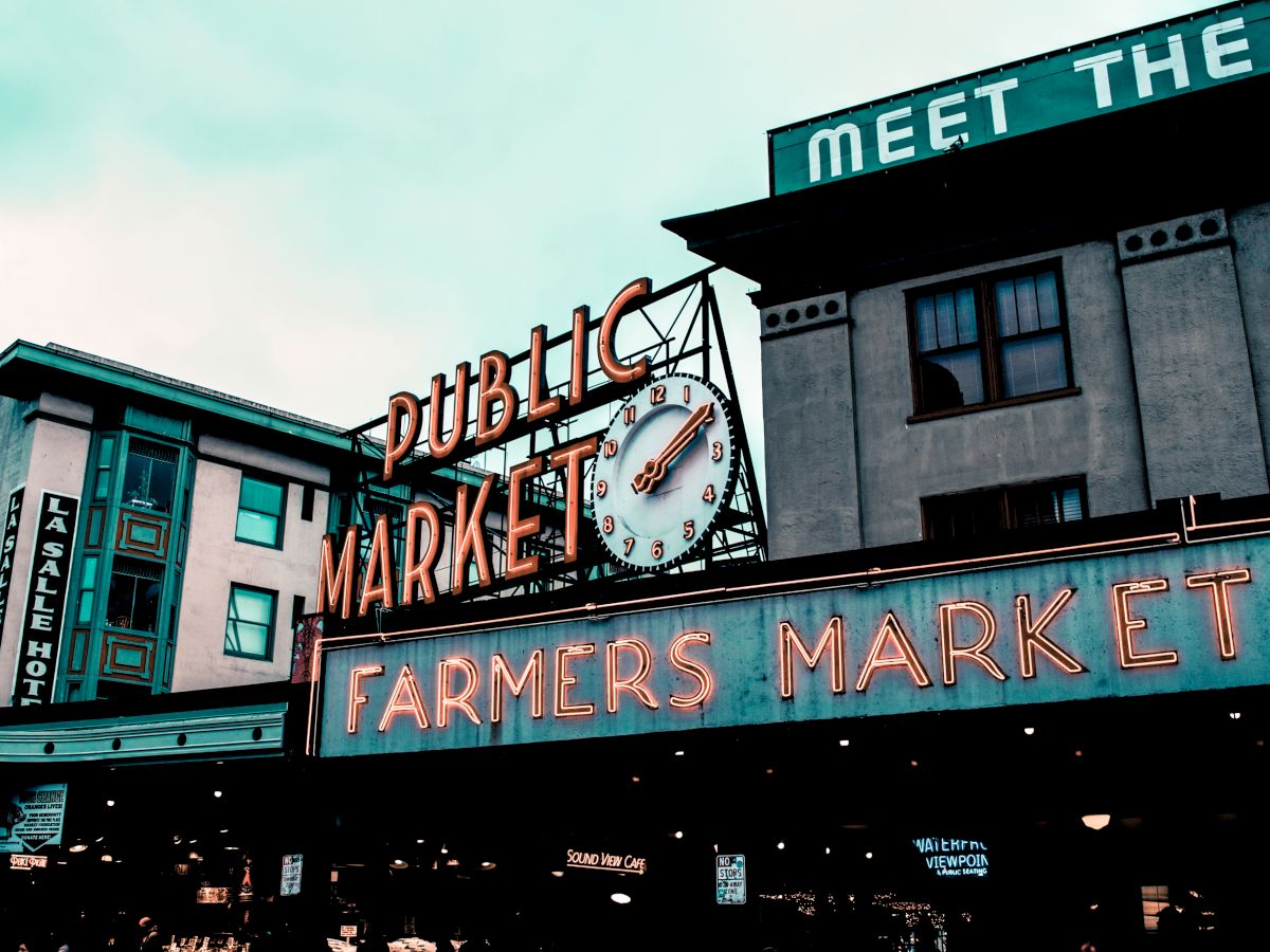 The image shows the neon signage of a building with "Public Market" and "Farmers Market" on it, along with a clock and multiple storefronts visible.