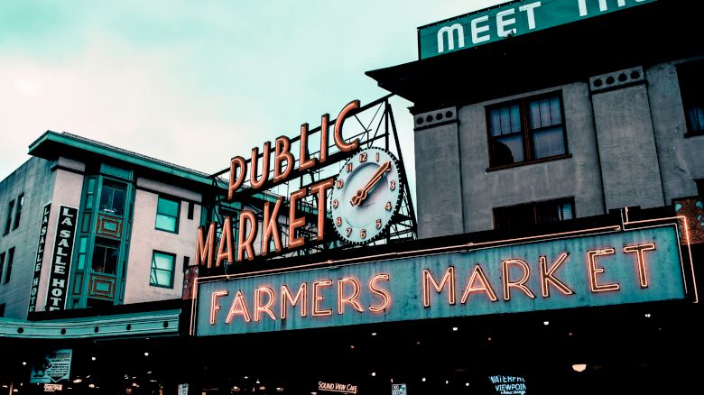 The image shows the neon signage of a building with "Public Market" and "Farmers Market" on it, along with a clock and multiple storefronts visible.