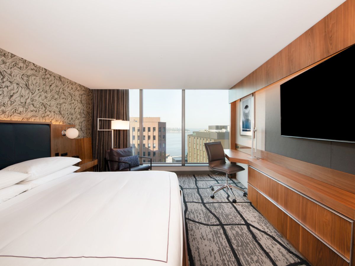 A modern hotel room with a large bed, desk, and chair, featuring a wall-mounted TV, large windows with a city view, and stylish decor ending the sentence.