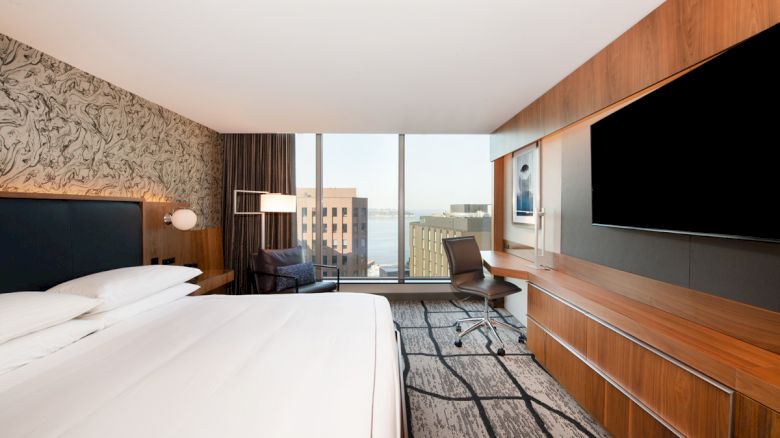 A modern hotel room with a large bed, desk, and chair, featuring a wall-mounted TV, large windows with a city view, and stylish decor ending the sentence.