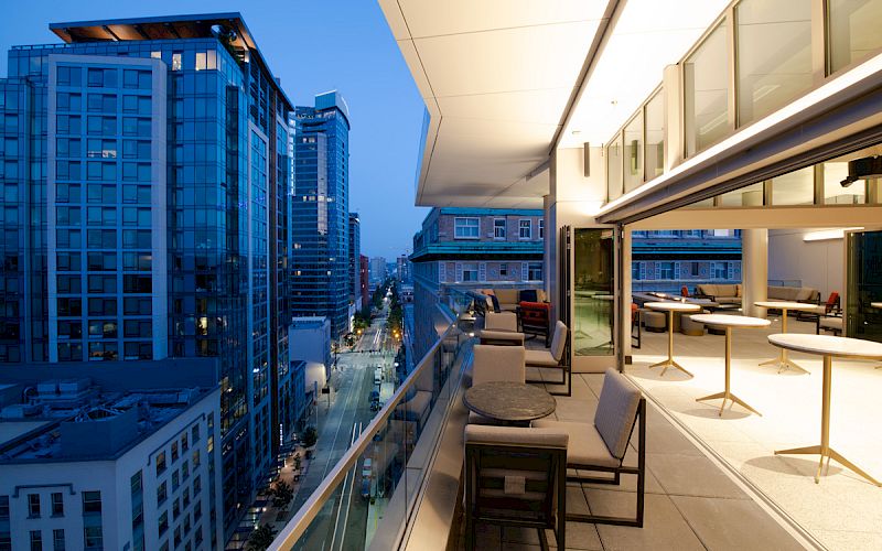 A modern cityscape view at dusk from a balcony, featuring tall buildings, a stylish outdoor seating area, and lit street below the balcony.