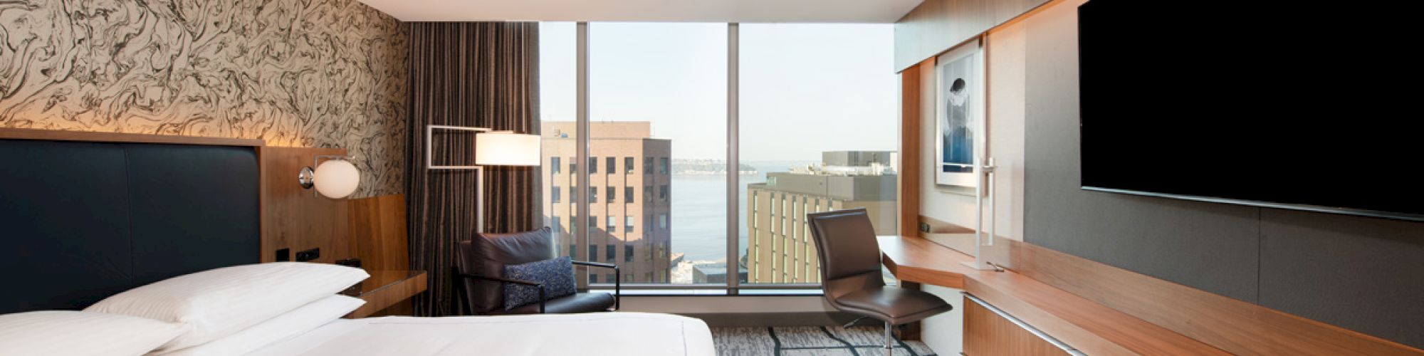 This image shows a modern hotel room with a king-sized bed, a large TV, a desk and chair, and a window offering a city and water view.