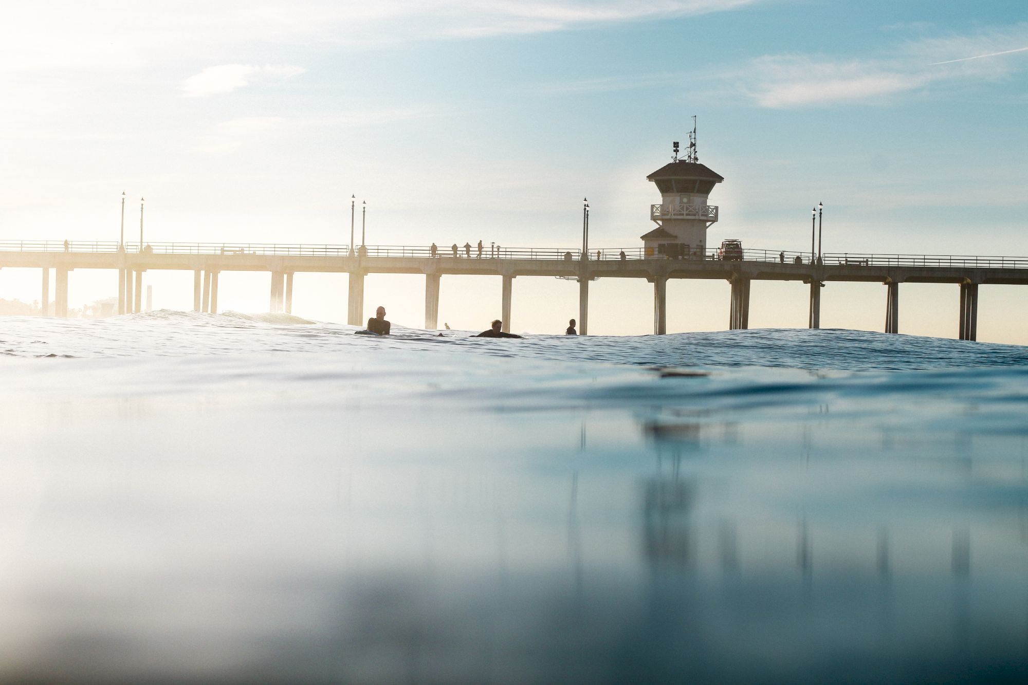 A pier extends out over calm waters with people walking on it and a lifeguard tower at the end. The scene is serene and bathed in sunlight.